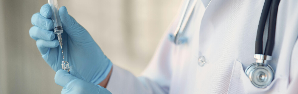 Ambulante Zusatzversicherungen Ein Arzt trägt hellblaue medizinische Einweghandschuhe und hält eine Spritze mit einer Nadel, die er mit einer Flüssigkeit aus einer Flasche füllt. Ein Stethoskop hängt um seinen Hals.