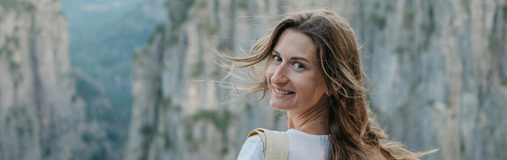 Betriebliche Altersversorgung Frau mit langen Haaren, die ins Bild lächelt; im Hintergrund sind steile, felsige Klippen und Natur zu sehen.
