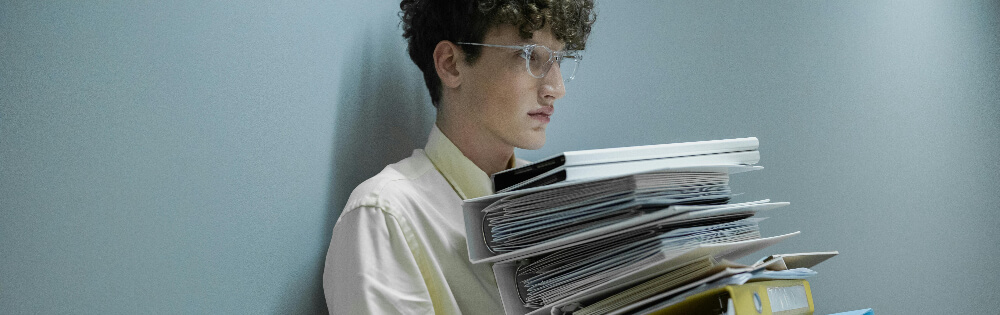 Versicherungsschutz für Schüler und Praktikanten Ein junger Mann mit Locken und Brille steht an einer grauen Wand und hält einen großen Stapel Dokumente und Ordner in den Armen.