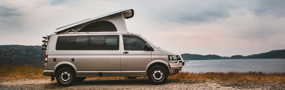 Wohnmobil & Camper Silberner Campervan mit hochgeklapptem Dach steht an einem Seeufer mit bewölktem Himmel und Hügeln im Hintergrund.