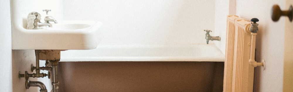 Leitungswasser Ein kleines Badezimmer mit einer freistehenden Badewanne, einem Waschbecken mit alten Wasserhähnen und einem Heizkörper an der Wand. Die Sanitäranlagen wirken altmodisch und der Raum ist mit weißen Wänden ausgestattet.