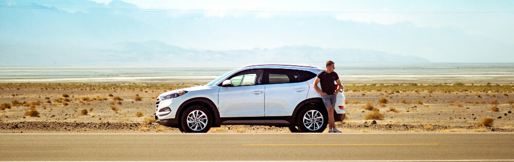 Mietwagenschutz Ein Mann steht neben einem silbernen SUV am Straßenrand in einer trockenen, wüstenartigen Landschaft. Im Hintergrund sind Berge und ein blauer Himmel zu sehen.