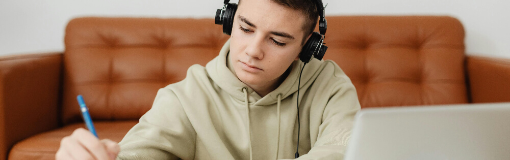 Einkommensschutz Ein junger Mann sitzt auf einem Sofa und trägt Kopfhörer. Er konzentriert sich auf das Schreiben mit einem Stift auf Papier, während vor ihm ein Laptop steht.